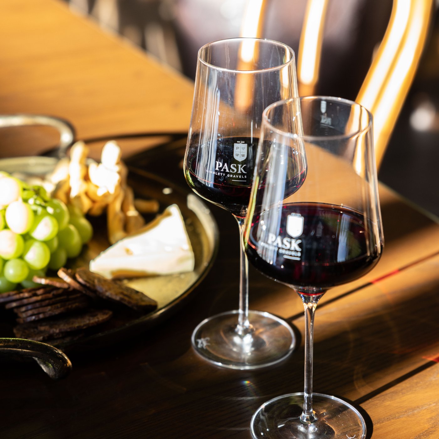 Two Pask branded red wine glasses on a wooden table beside a cheese and grape platter