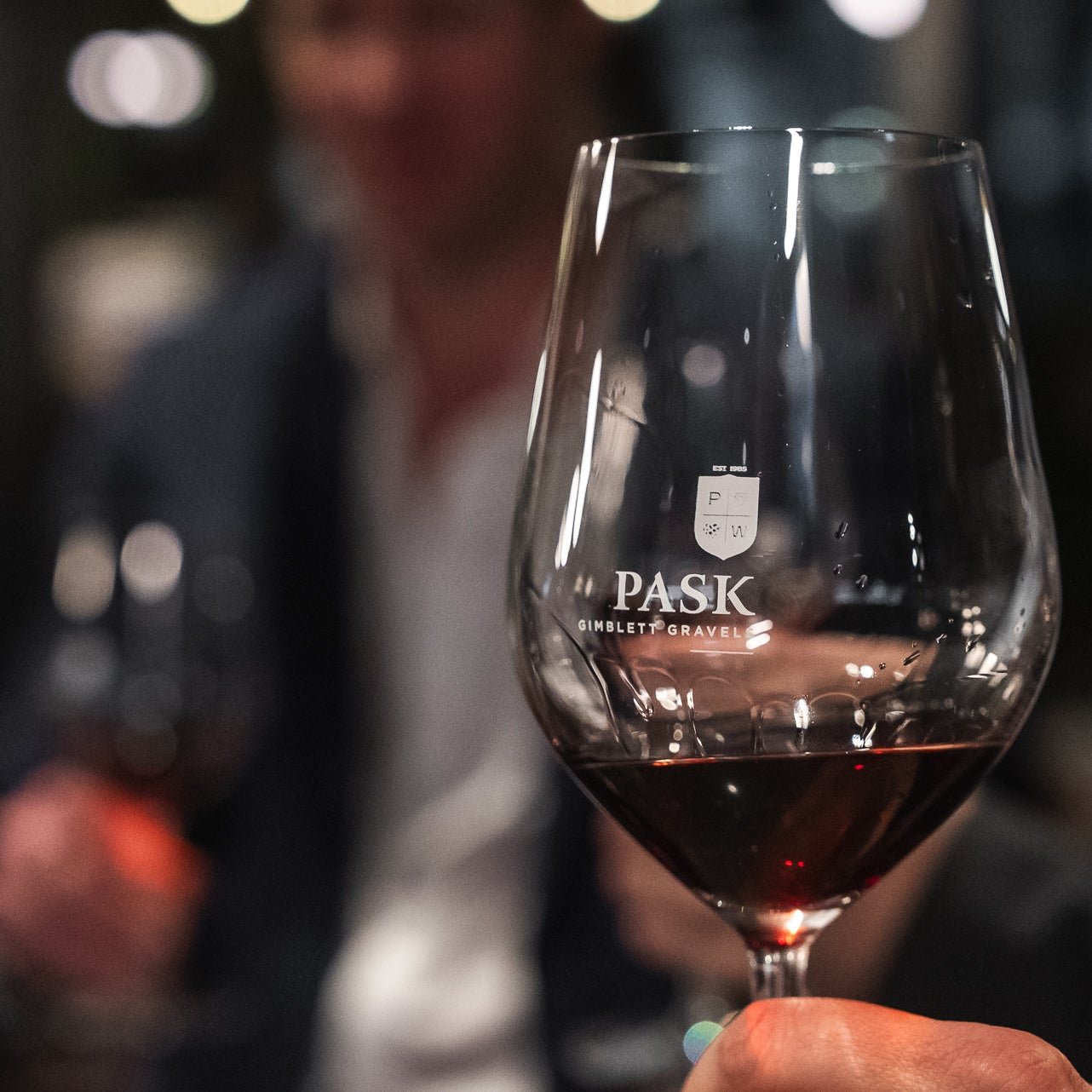 A hand holding a Pask Gimblett Gravel branded wine glass with red wine, person in background at a dimly lit event.