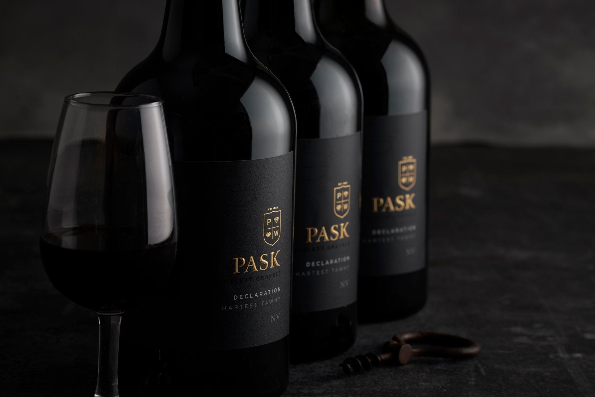 Three bottles of Pask Declaration red wine with a filled wine glass and corkscrew on a dark stone surface.