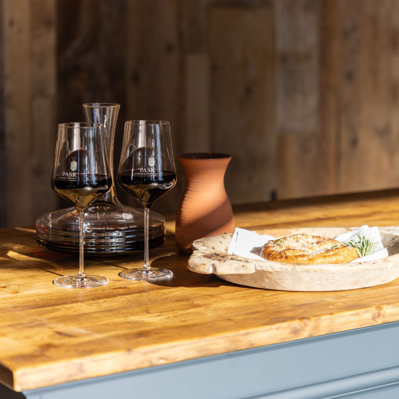 Two Pask branded wine glasses and a decanter beside a rustic wooden board with baked food on a timber counter