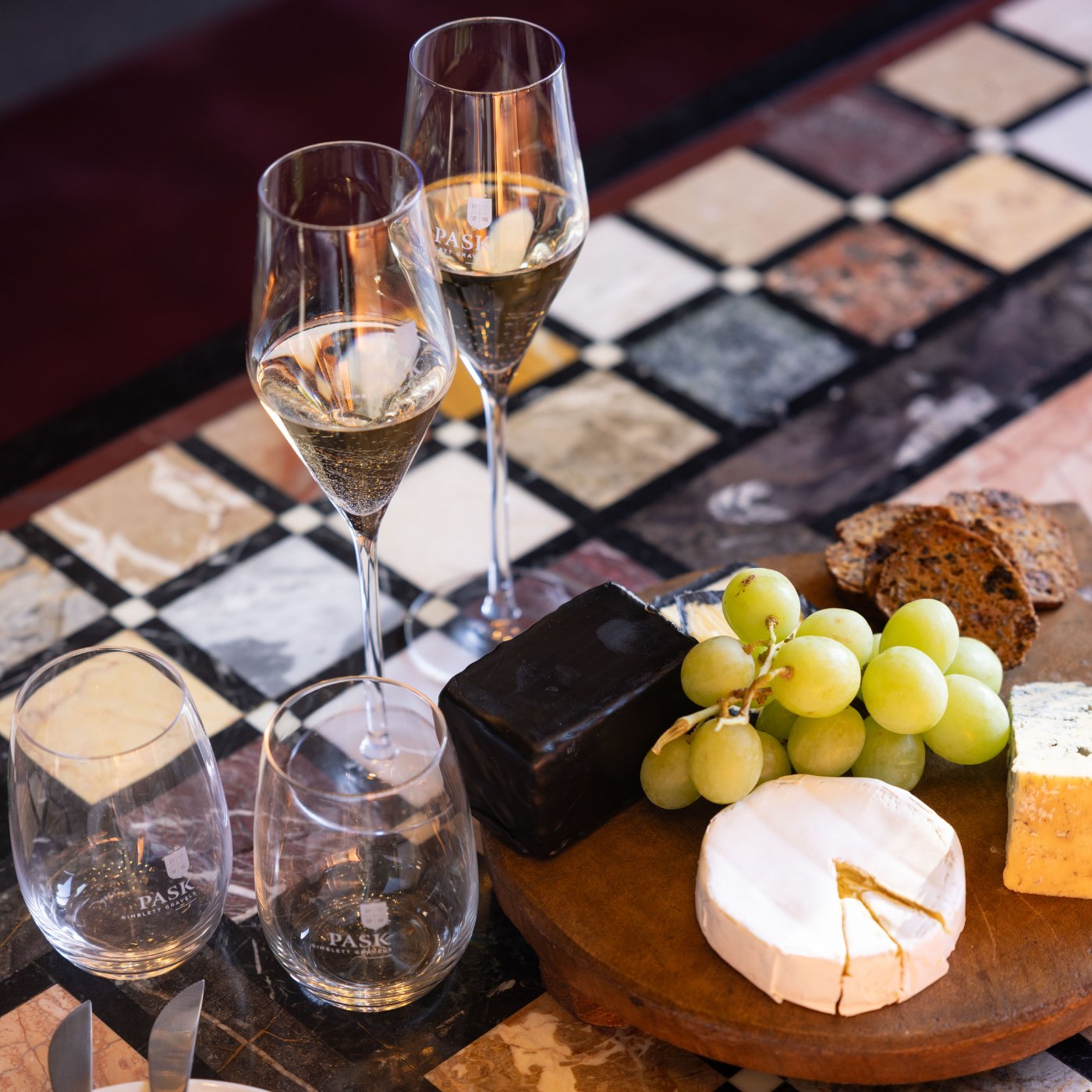 Pask branded wine glasses and two flutes of white wine alongside a cheese board with grapes, brie, and crackers