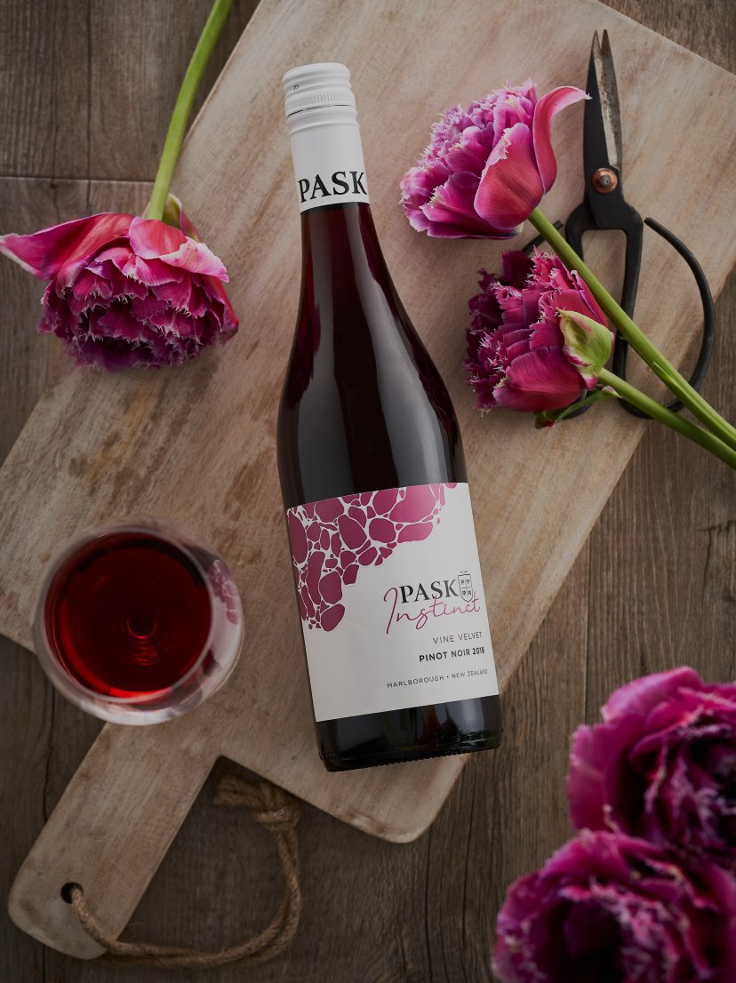 Pask Instinct Pinot Noir bottle on a wooden board with a glass of red wine, pink tulips, and garden scissors