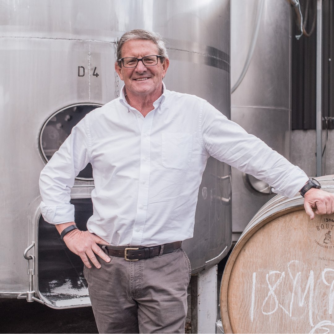 Man in white shirt leaning on an oak barrel in front of stainless steel winery tanks