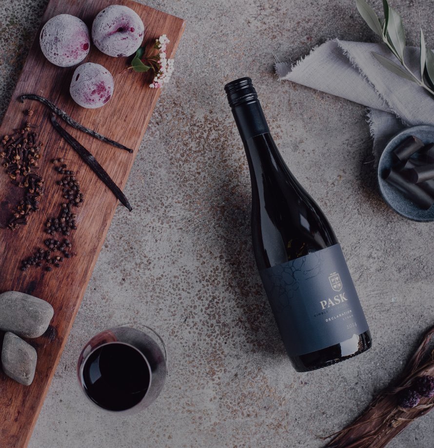 Pask Declaration red wine bottle laid flat beside a glass of red wine, plums, vanilla pods, and peppercorns on a stone surface