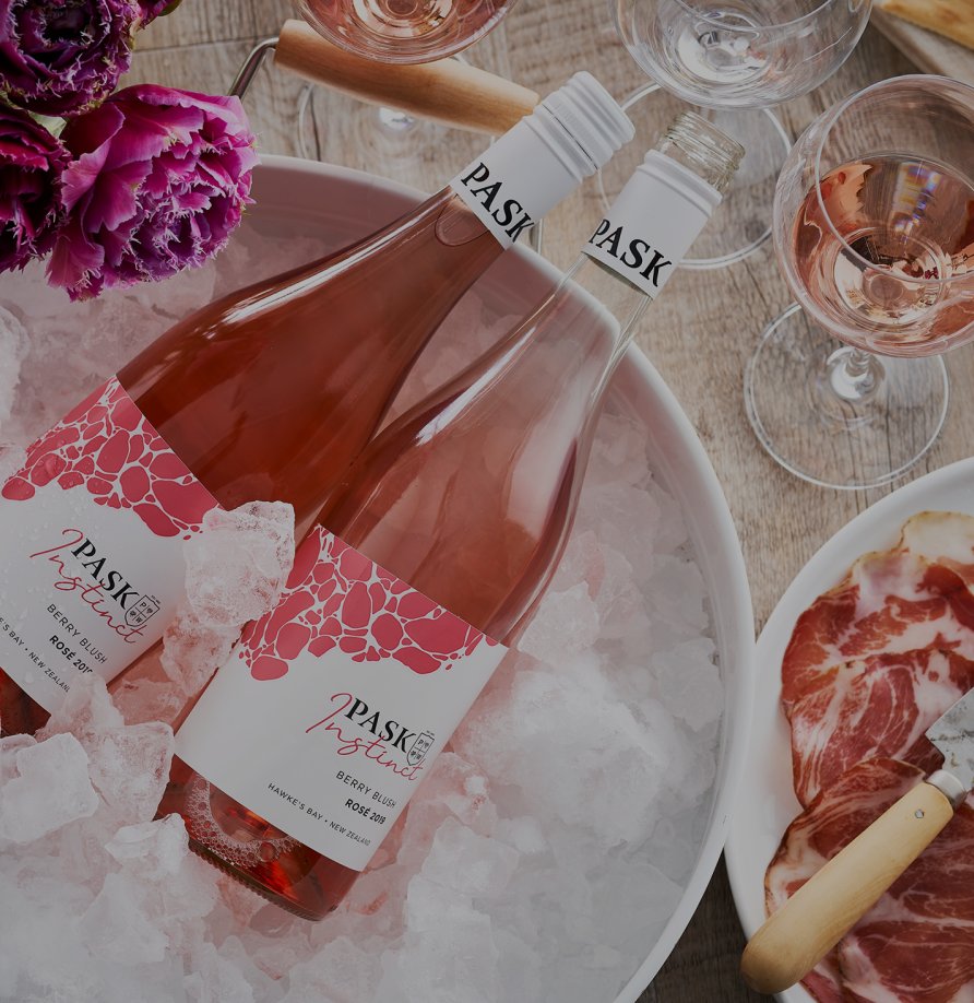 Two bottles of Pask Instinct Berry Blush Rosé chilling on ice, with wine glasses, cured meat, and pink flowers on a wooden table