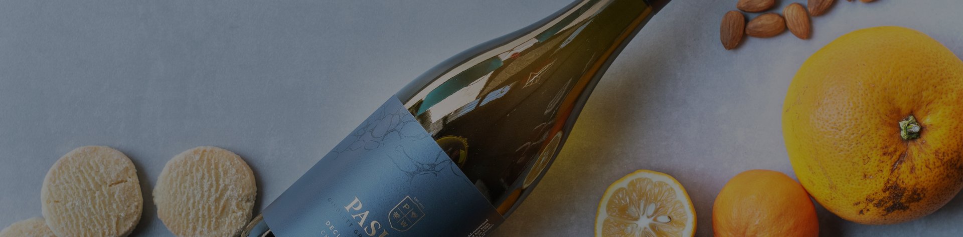 Pask wine bottle lying on its side beside citrus fruits and almonds on a light surface