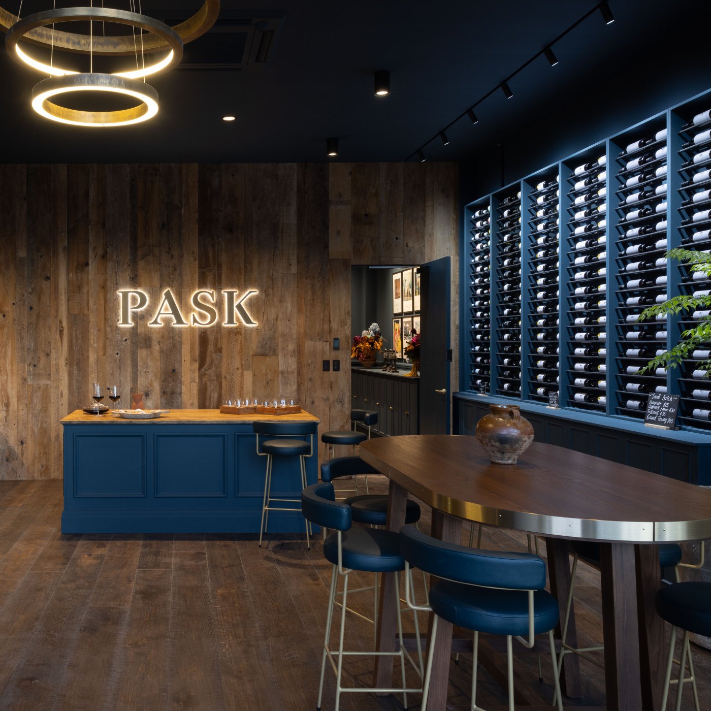 Pask Winery tasting room interior with illuminated PASK sign, navy tasting counter, bar stools, and floor-to-ceiling wine rack