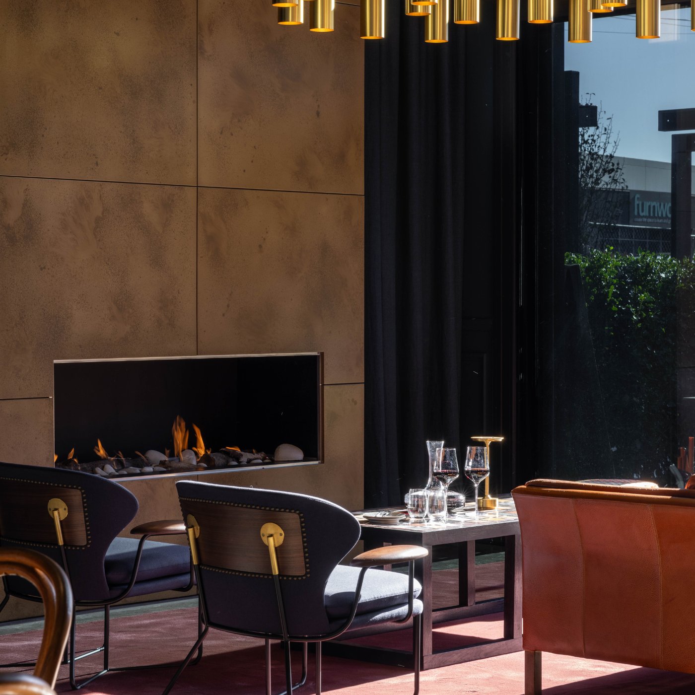 Elegant tasting room interior with a lit fireplace, dark upholstered chairs, wine glasses on a table, and gold pendant lighting.